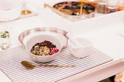 A styled tabletop scene with a white bowl filled with mixed seeds and dried herbs, a white mortar and pestle, a gold spoon, and a striped placemat on a white table.