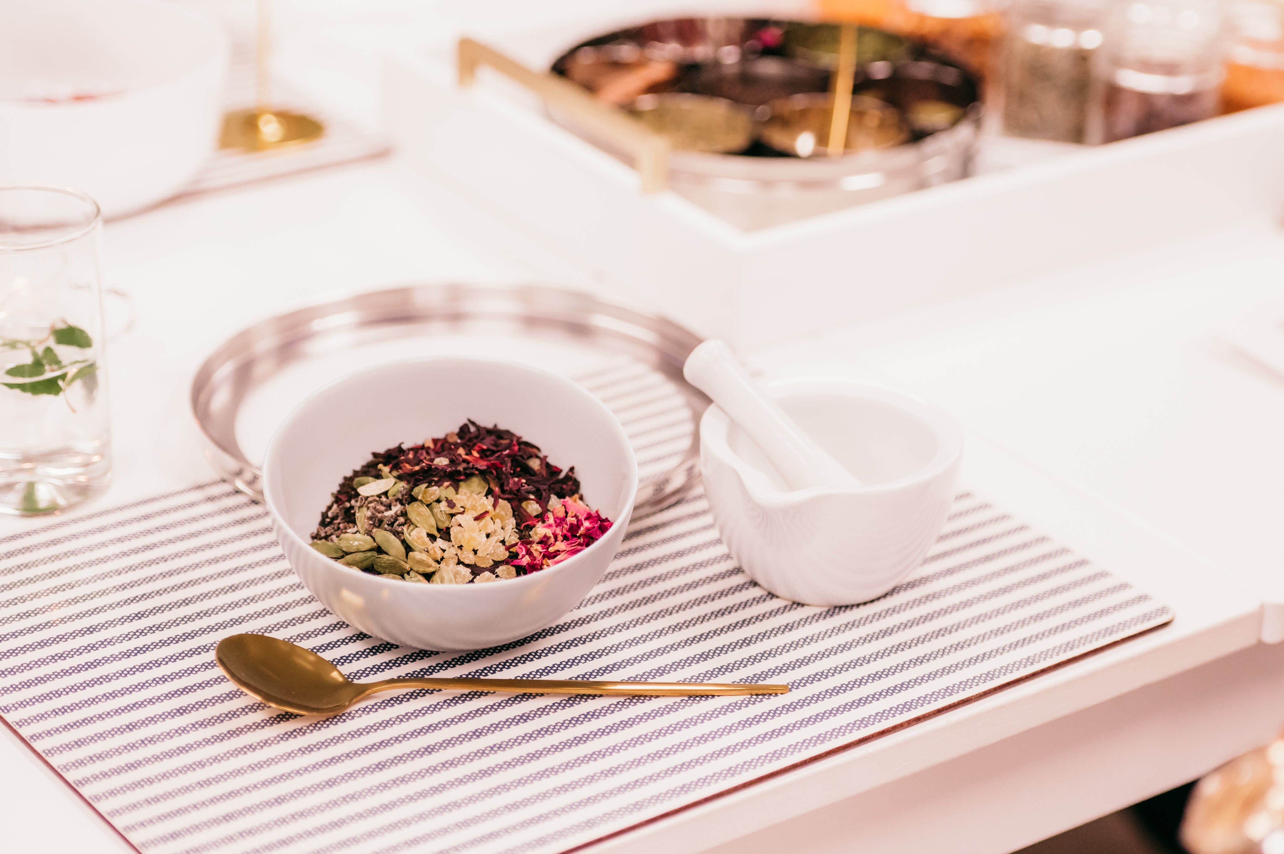 A styled tabletop scene with a white bowl filled with mixed seeds and dried herbs, a white mortar and pestle, a gold spoon, and a striped placemat on a white table.