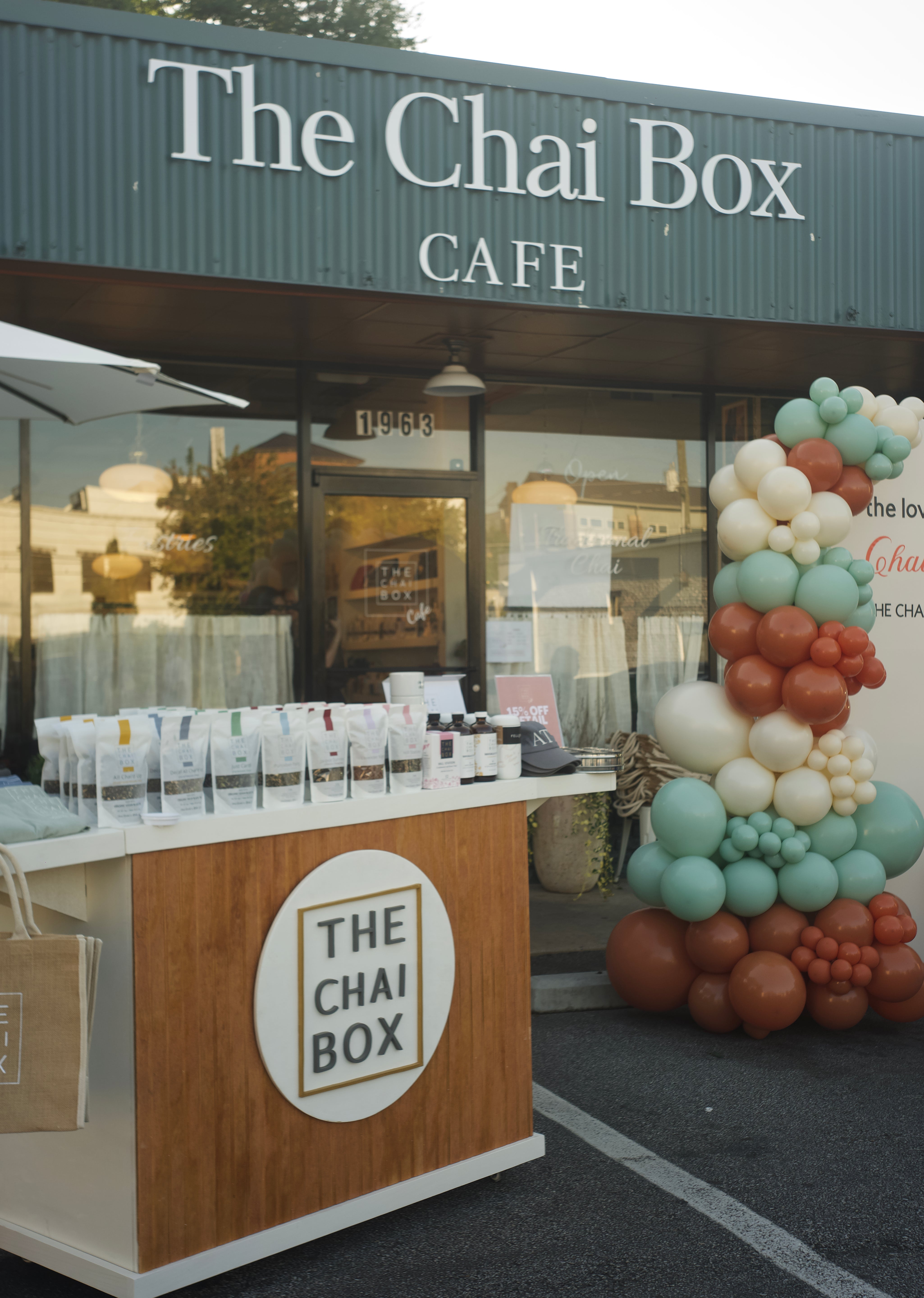 The Chai Box Cafe storefront with a branded wooden counter and a large balloon arch in teal, cream, and terracotta beside the entrance.
